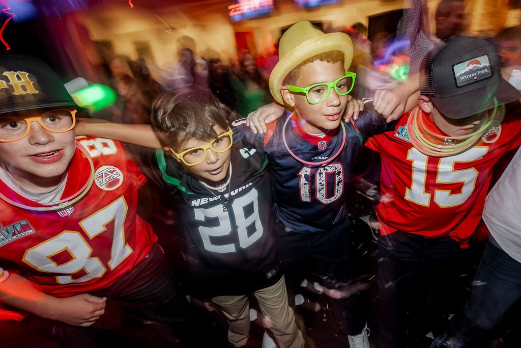 Four boys wearing sports jerseys and glasses, dancing together at a lively indoor party.