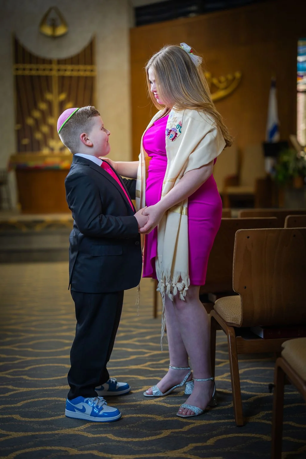 A boy and woman holding hands and looking at each other inside a room with wooden walls and chairs.