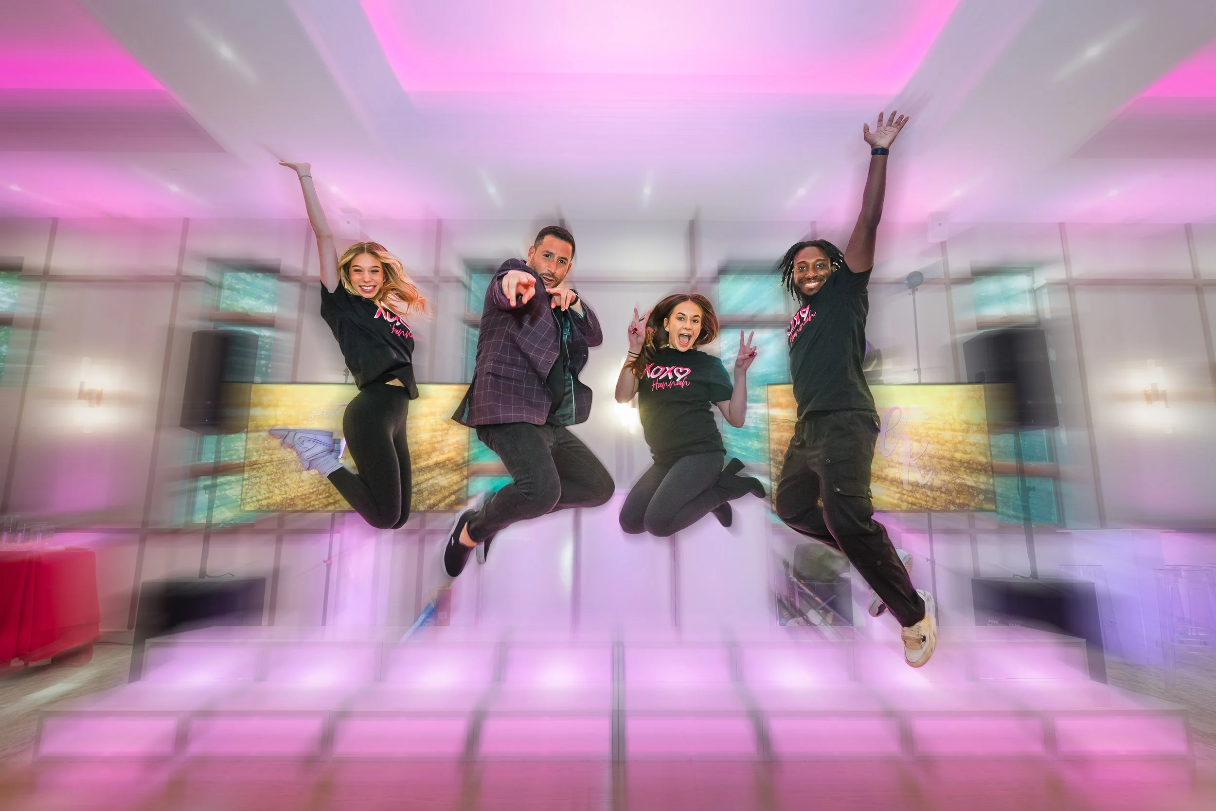 Four people jumping in the air indoors at a party or celebration with colorful lighting and screens in the background.