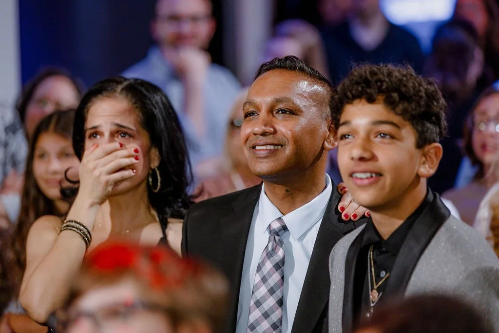 A diverse group of people at a celebration or event, with a woman covering her mouth and a man and young boy smiling, surrounded by other attendees in the background.