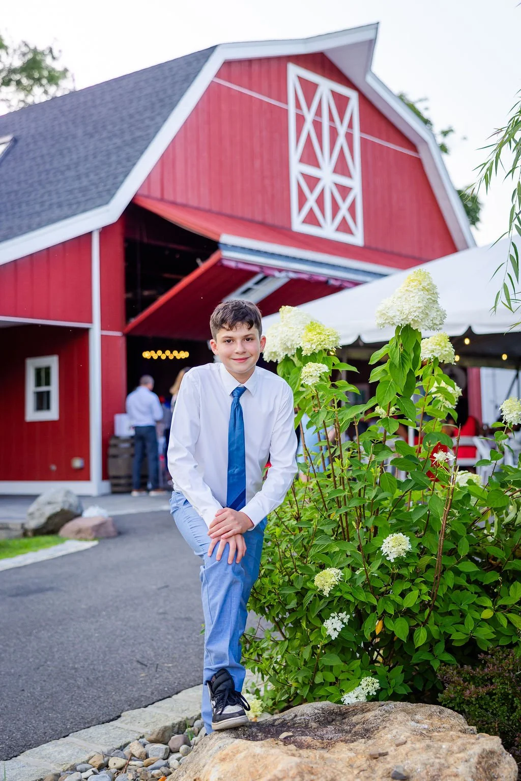 A young boy in a white shirt, blue tie, and light blue pants posing outdoors near a bush with white flowers, in front of a large red barn with white trim, under a white canopy, during daytime.