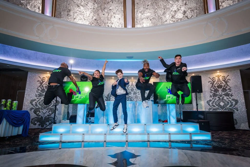 Group of five people jumping in the air on a stage at an event, with bright lights and digital screens in the background.