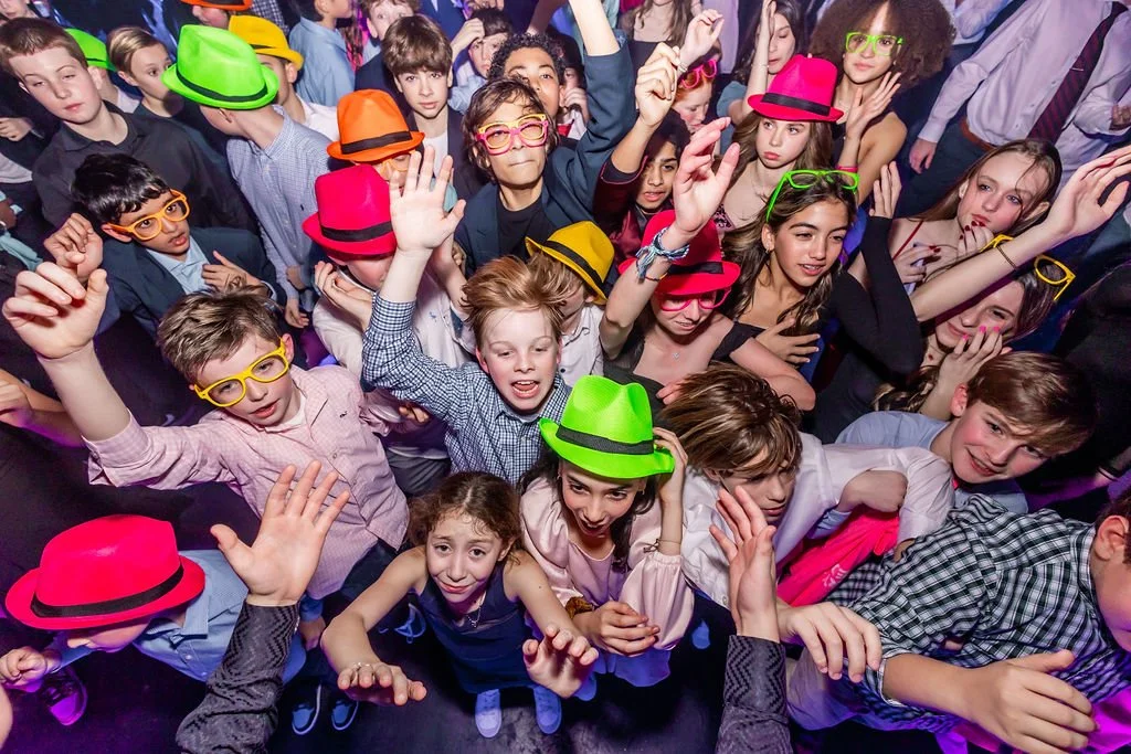 A large group of children at a party or event, wearing colorful hats and glasses, dancing and having fun.