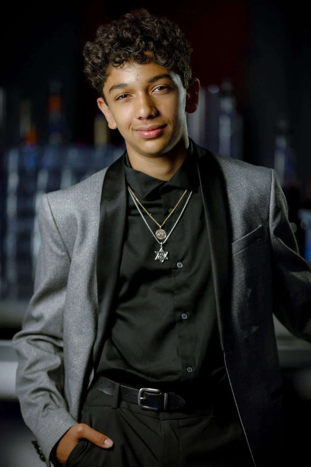 Young man with curly hair in a black shirt and gray blazer, wearing layered necklaces, standing confidently indoors.