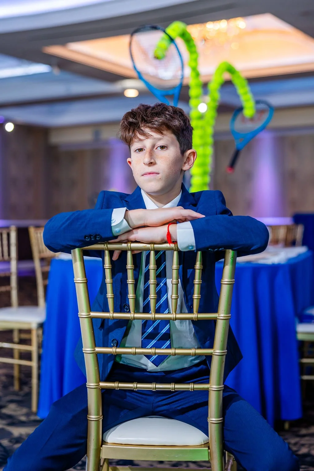 A young man in a blue suit and tie sitting at a table with his arms crossed over a gold-colored chair back, in a decorated event space with tennis rackets and fuzzy decorations hanging from the ceiling.