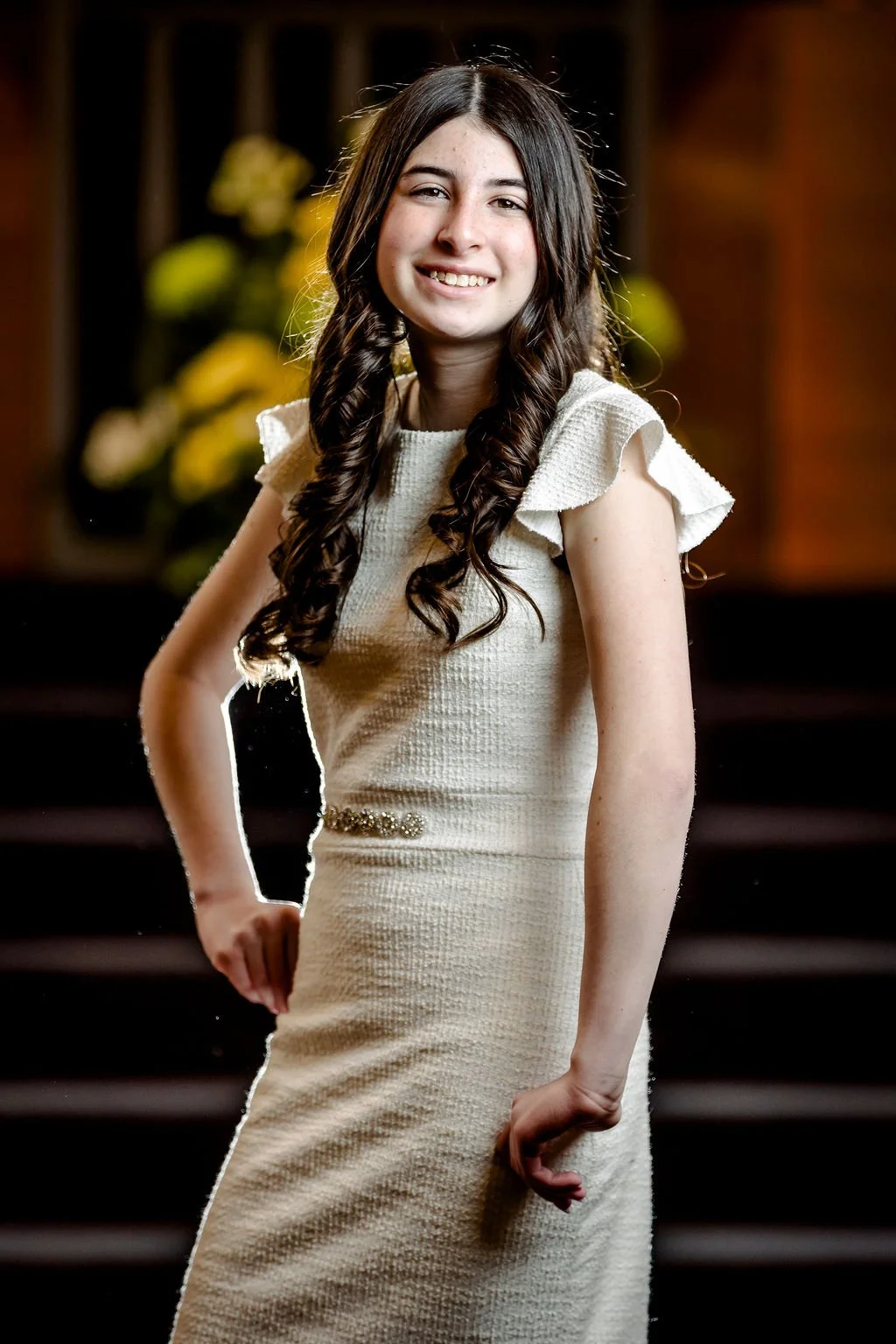 Young woman with long, curly hair wearing a cream-colored dress with ruffled sleeves, standing indoors with a blurred background of yellow flowers and wooden steps.