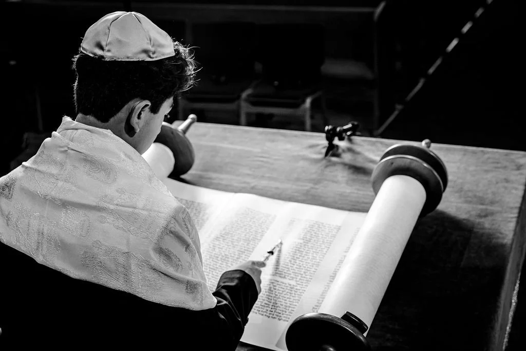 A young boy dressed in religious attire, reading from a large scroll on a wooden table, with a small rifle on the table.