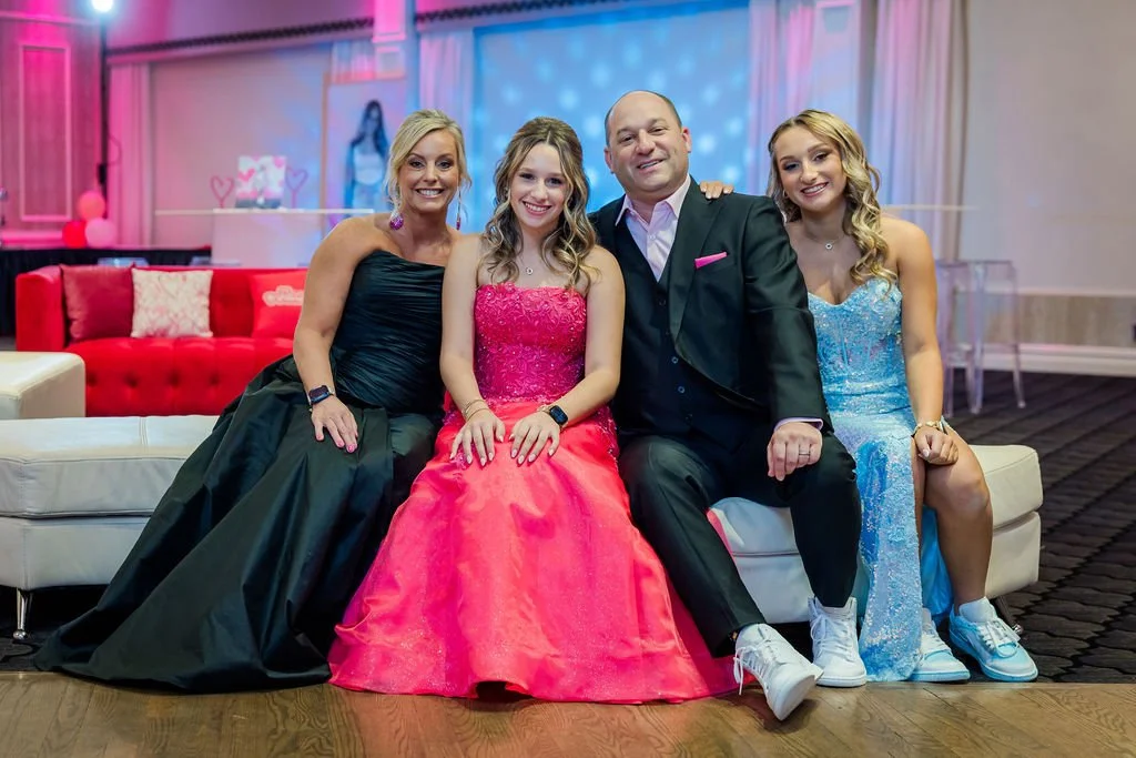 Four people sitting on a couch at a party or celebration, with colorful lighting and decorations in the background. The group includes two women in formal dresses, one young girl in a pink dress, and a man in a black suit.