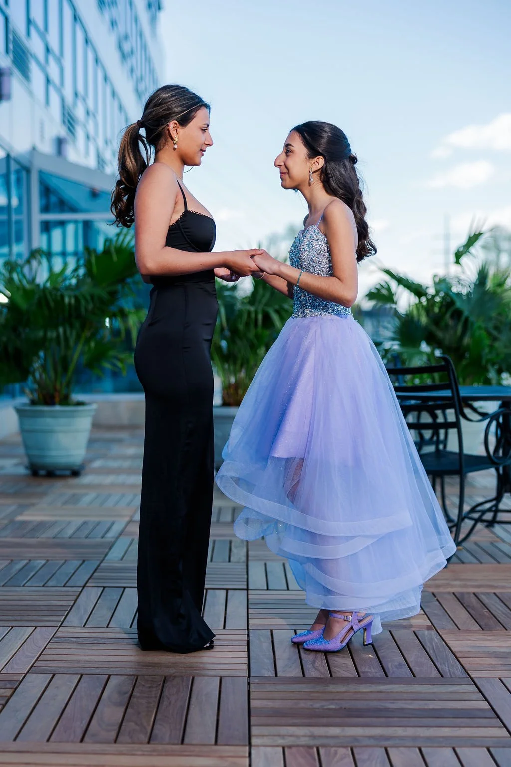 Two women holding hands and smiling at each other on an outdoor wooden deck, with large potted plants and a modern glass building in the background. One woman is wearing a black dress, and the other is in a sparkly top with a layered lavender tulle s