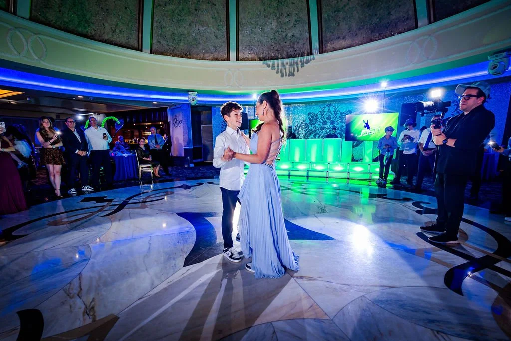 A young couple dancing in the center of a decorated ballroom with onlookers and a cameraman capturing the moment, illuminated by colorful lights.