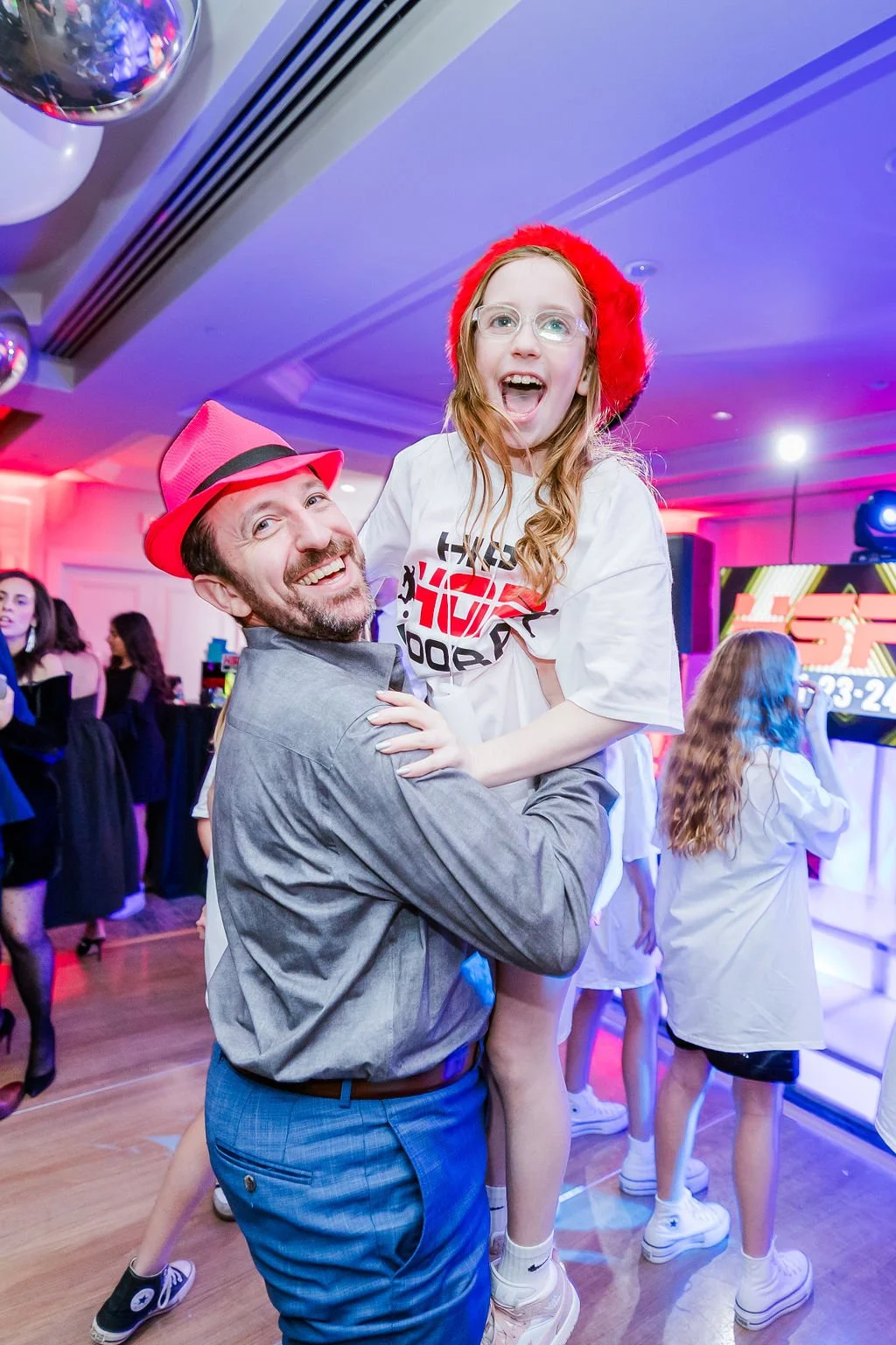 A man in a gray shirt and blue pants, wearing a pink hat, holding a young girl with red hair, glasses, and a red hat, at a festive indoor event with colorful lighting and other people in the background.