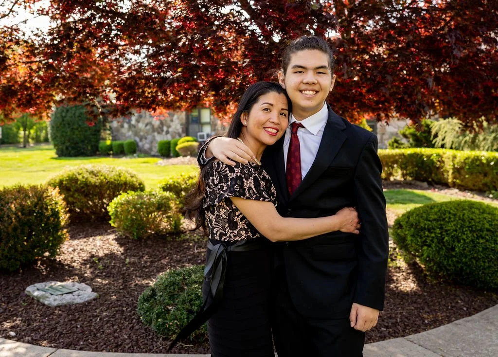 A woman and a young man hugging outdoors in a garden with bushes, trees, and a stone house in the background.