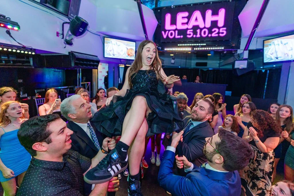 A young girl in a black dress being lifted on a celebration stage with people clapping and cheering around her, with a digital sign displaying "LEAH VOL. 5.10.25" in the background.
