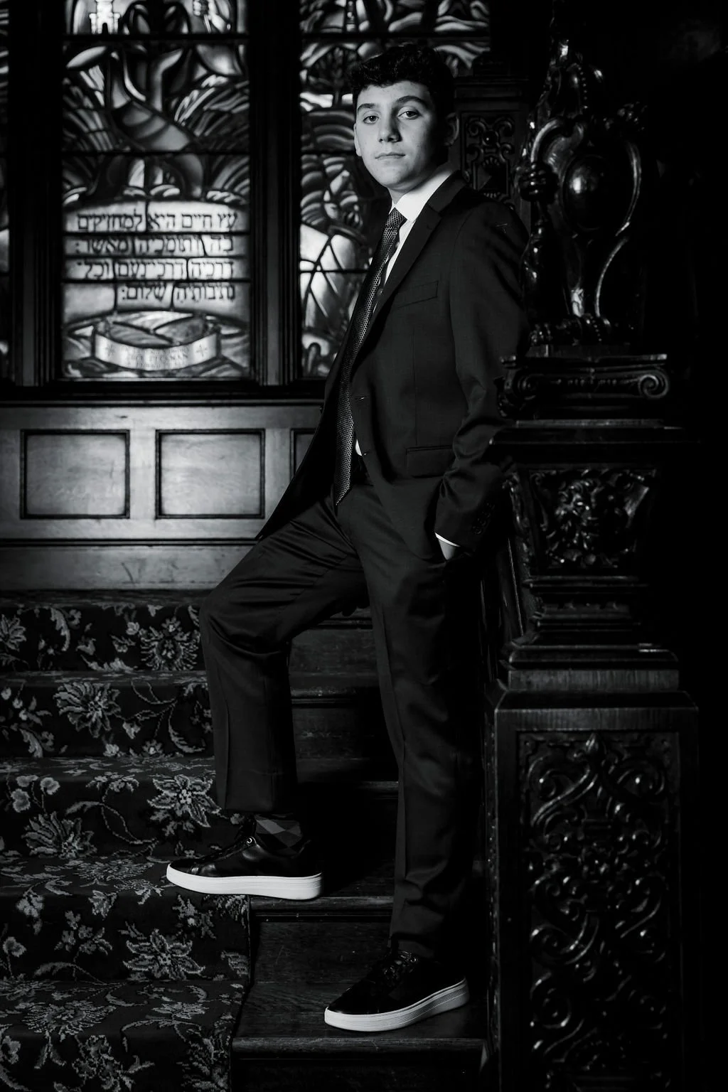 A young man in a suit standing on a staircase with ornate woodwork, black shirt, and sneakers in a formal setting with a decorative stained glass window behind him.