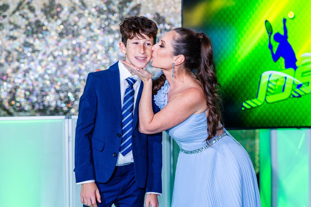 A woman in a light blue gown kisses a young boy in a blue suit on the cheek, with an animated basketball player on a screen in the background.