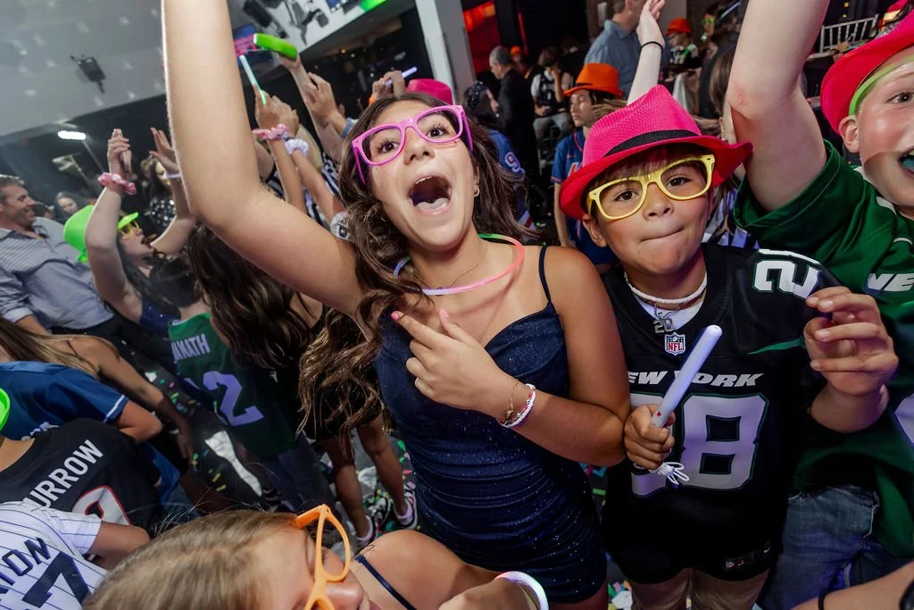 Children wearing sports jerseys and colorful glasses celebrating at a crowded party or stadium event.