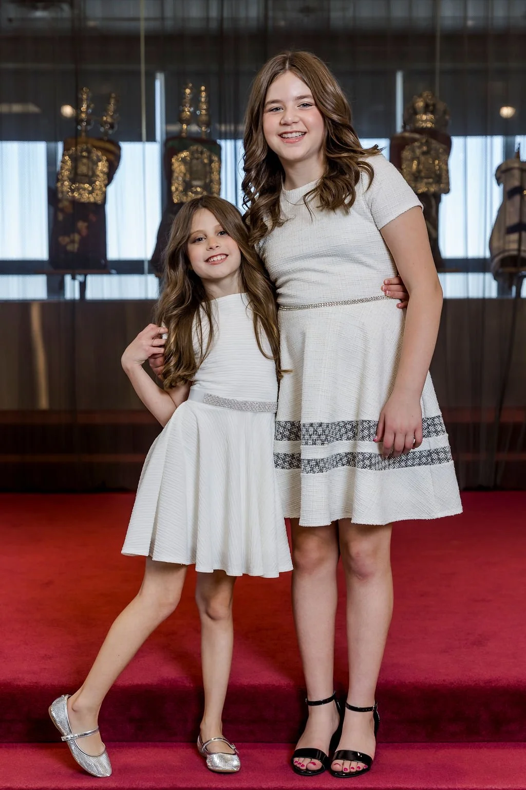 Two girls in white dresses standing on a red carpet in front of display cases with mannequins dressed in traditional clothing.