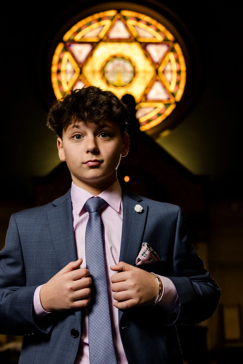 Young man in a suit and tie standing indoors with a stained glass window in the background.