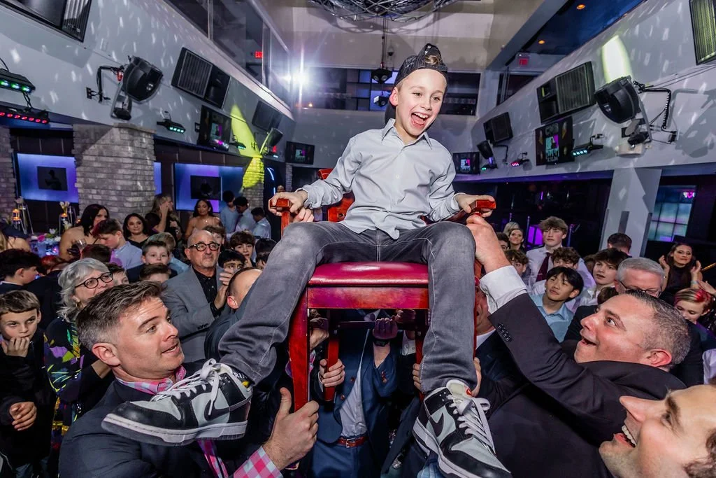 A young boy sitting on a chair held up by adults at a lively party or celebration with many people around in a decorated venue.