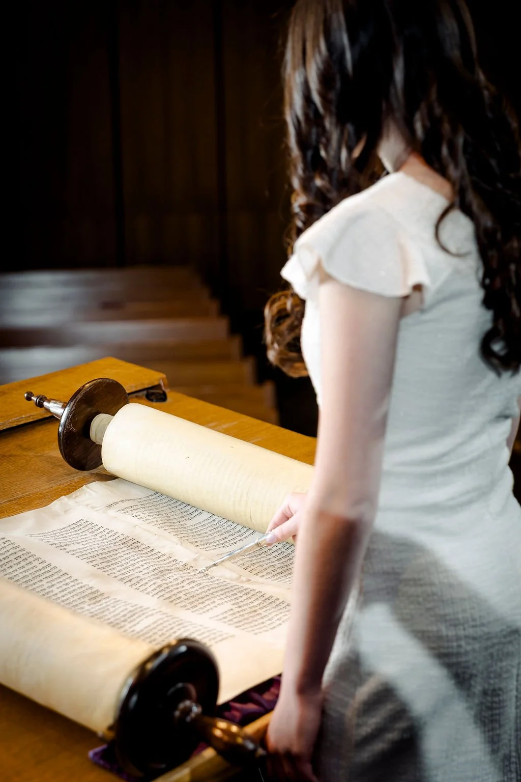 A woman with long, curly dark hair wearing a light-colored dress appears to be copying or examining an old handwritten document on a large scroll with a magnifying glass, on a wooden table.