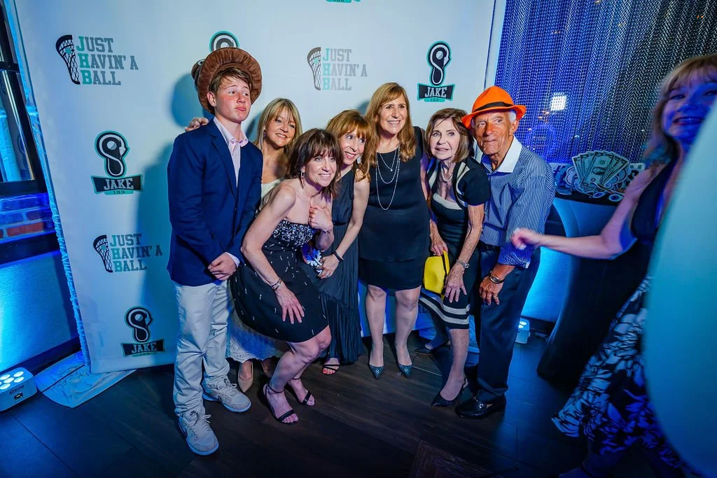 Group of seven people posing for a photo at an event with a backdrop showing the phrase 'Just Havin' a Ball' and a logo for Jake. The group includes a young man, four women, an older man, and another woman on the side. They are dressed in semi-formal