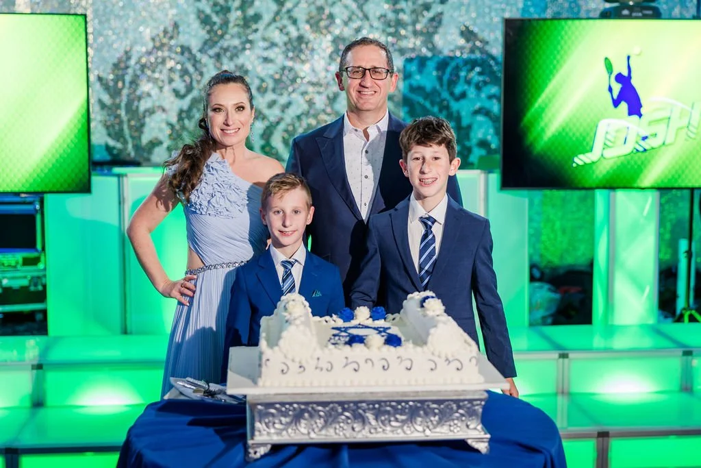 A family celebrating a birthday with three people, two boys and one woman, standing behind a table with a large birthday cake. A man is standing at the back, smiling. The background features green lighting and a screen with a tennis player graphic.
