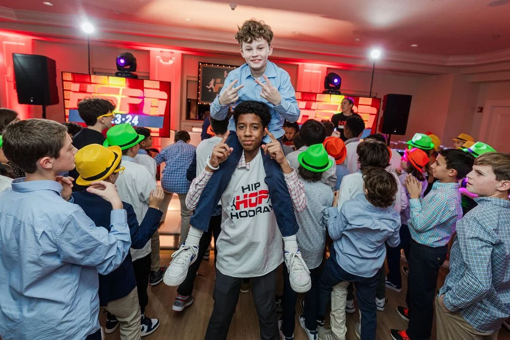 A group of children in a party room with colorful hats, dancing and enjoying a celebration with a DJ in the background, some wearing sunglasses, and a boy sitting on another boy's shoulders.