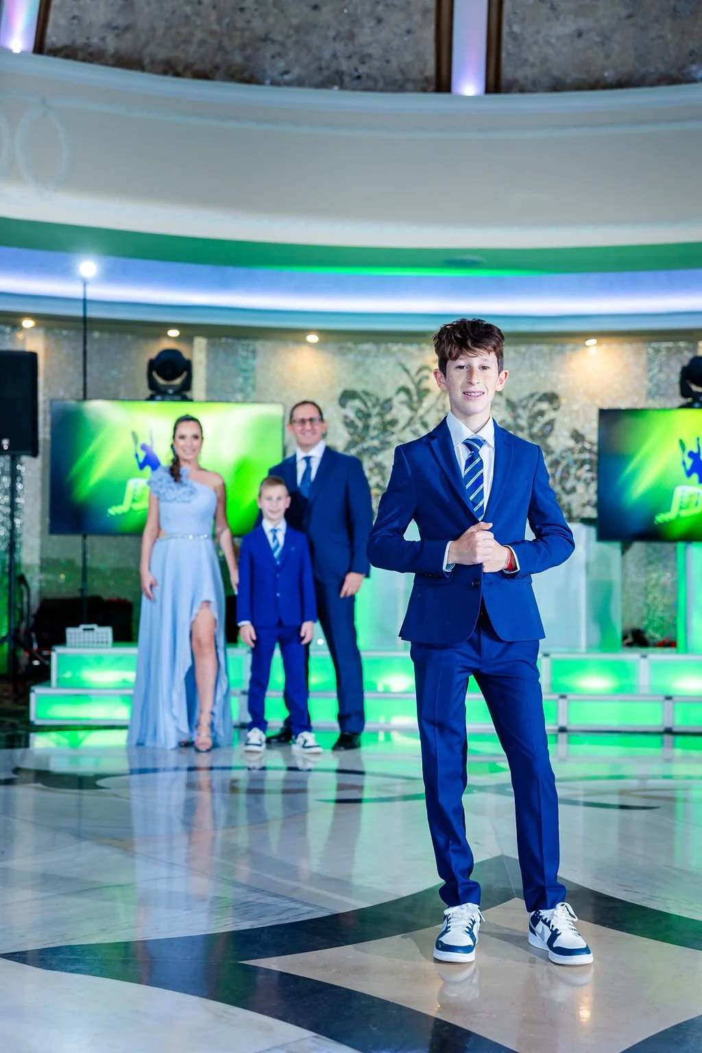 A young boy in a blue suit standing with hands clasped in front of a family at a social event, with a dance floor and large screens displaying graphics in the background.