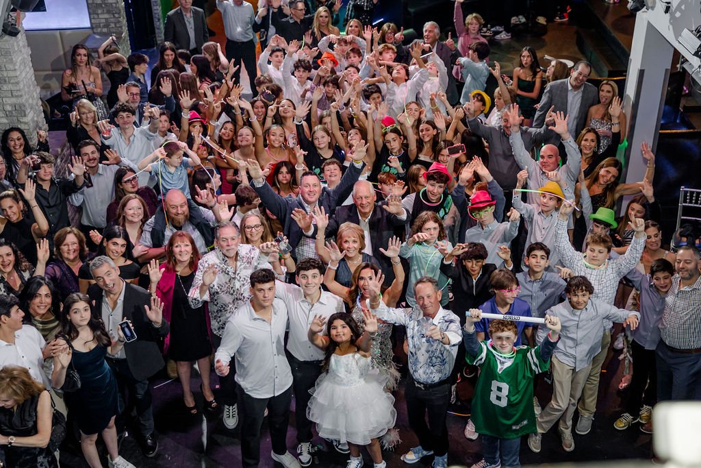 A large diverse group of people including children, teenagers, and adults gathered at an indoor event, smiling and raising their hands in celebration.