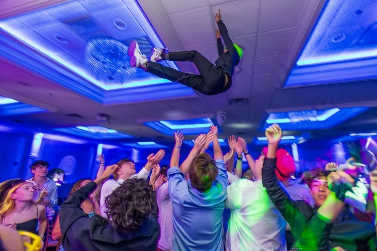 A group of young people dancing and celebrating at a party with blue and purple lighting, some with raised hands, while a person in black and green clothes is being lifted or thrown into the air.