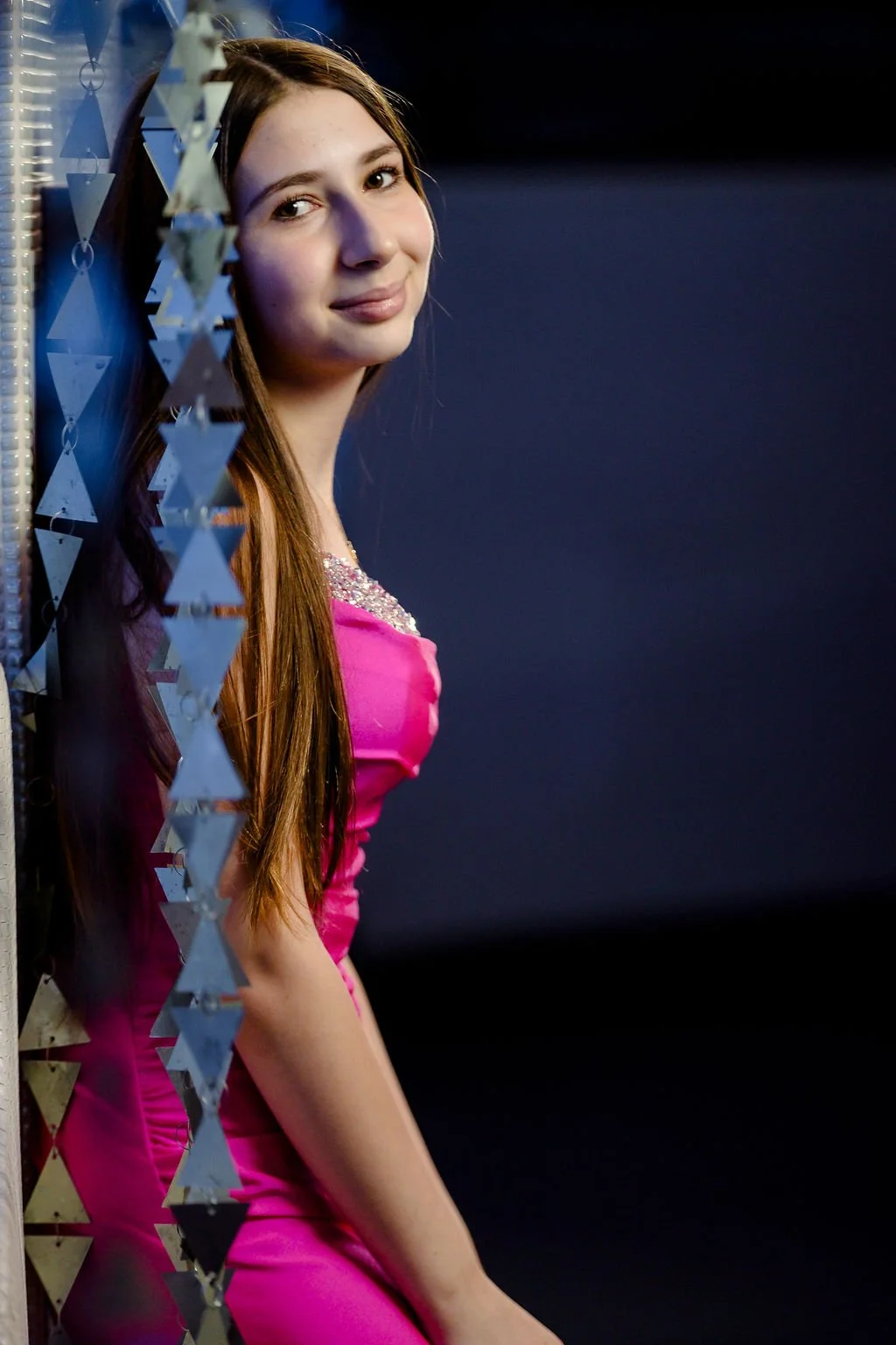 A young woman with long brown hair wearing a pink dress with embellishments on the neckline, smiling and leaning against a reflective surface with hanging silver triangular decorations on one side and a dark, plain background.