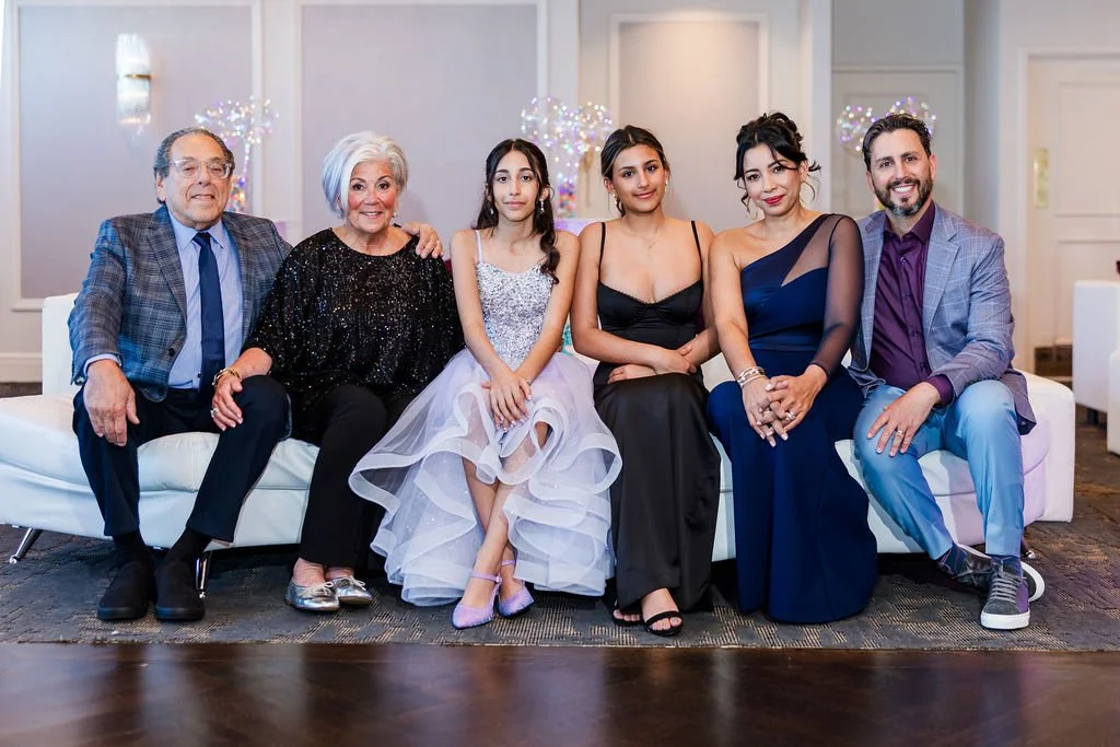Six people sitting on a white sofa in a well-lit room, dressed in formal attire, smiling for a group photo.