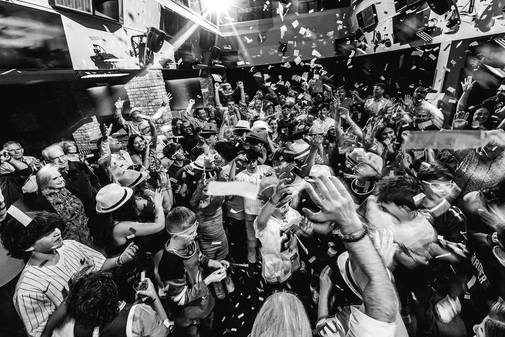 Crowd of people celebrating, dancing, and throwing confetti at a lively indoor party or event.