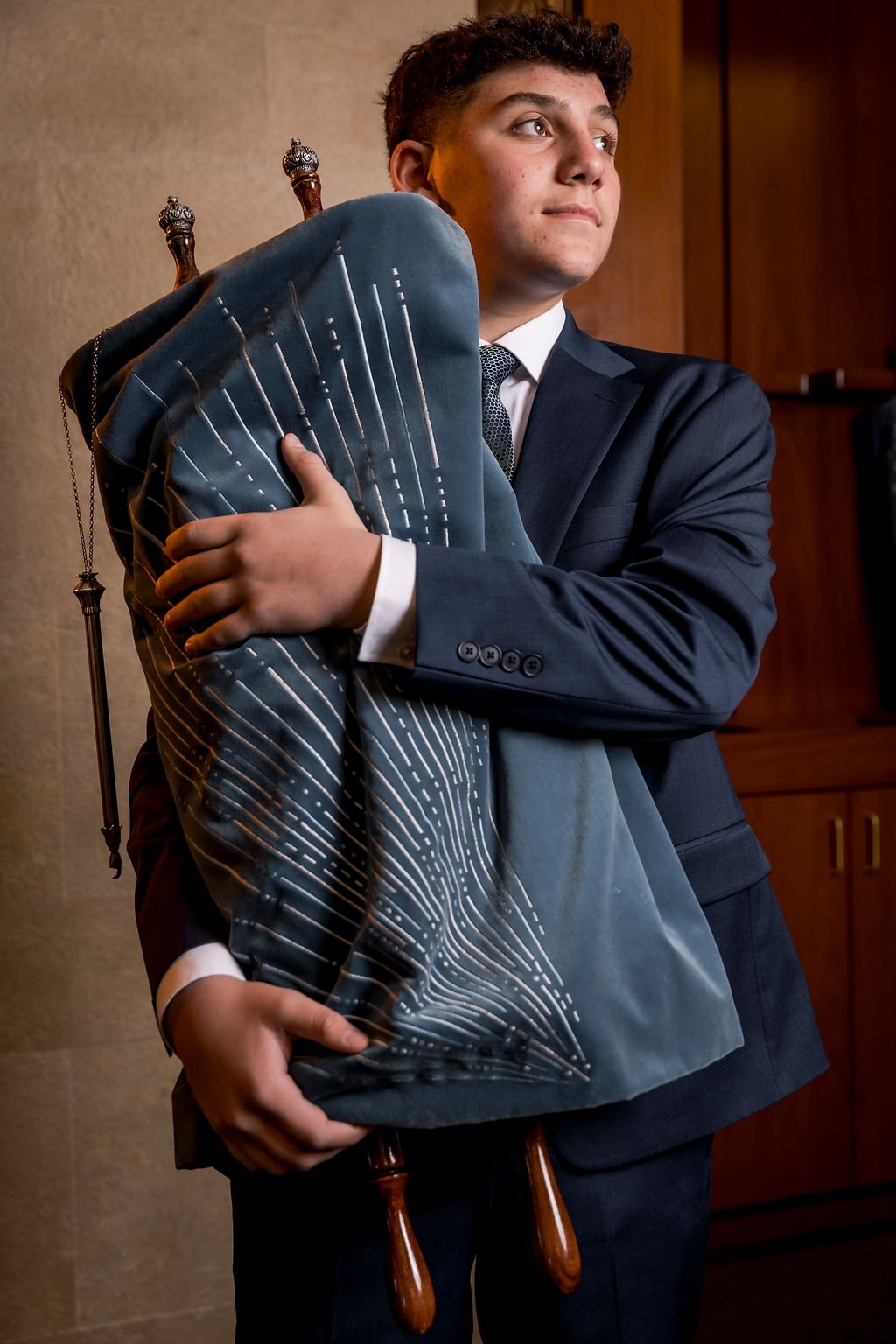 A young man in a suit holding a chair with a modern design, standing indoors near a wooden cabinet.