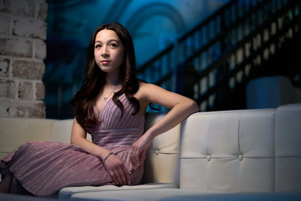 A young woman with long dark hair, wearing a sparkly pink dress, sitting on a cream-colored sofa in a dimly lit environment with a modern, industrial background.
