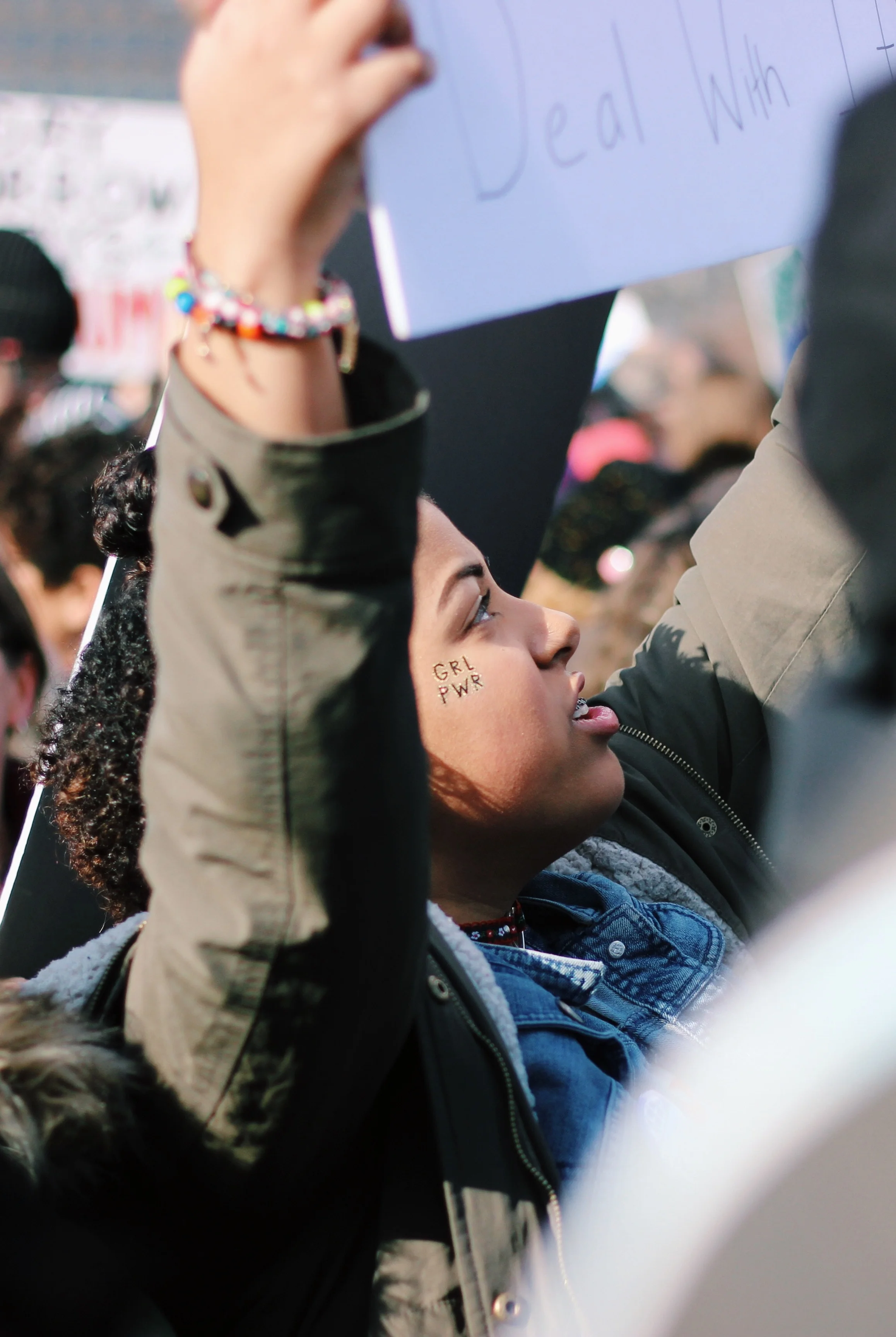 A younger person with brown skin and braces holding their arms up at a gathering of people, they have a temporary tattoo on their cheek that says “GRL PWR”.