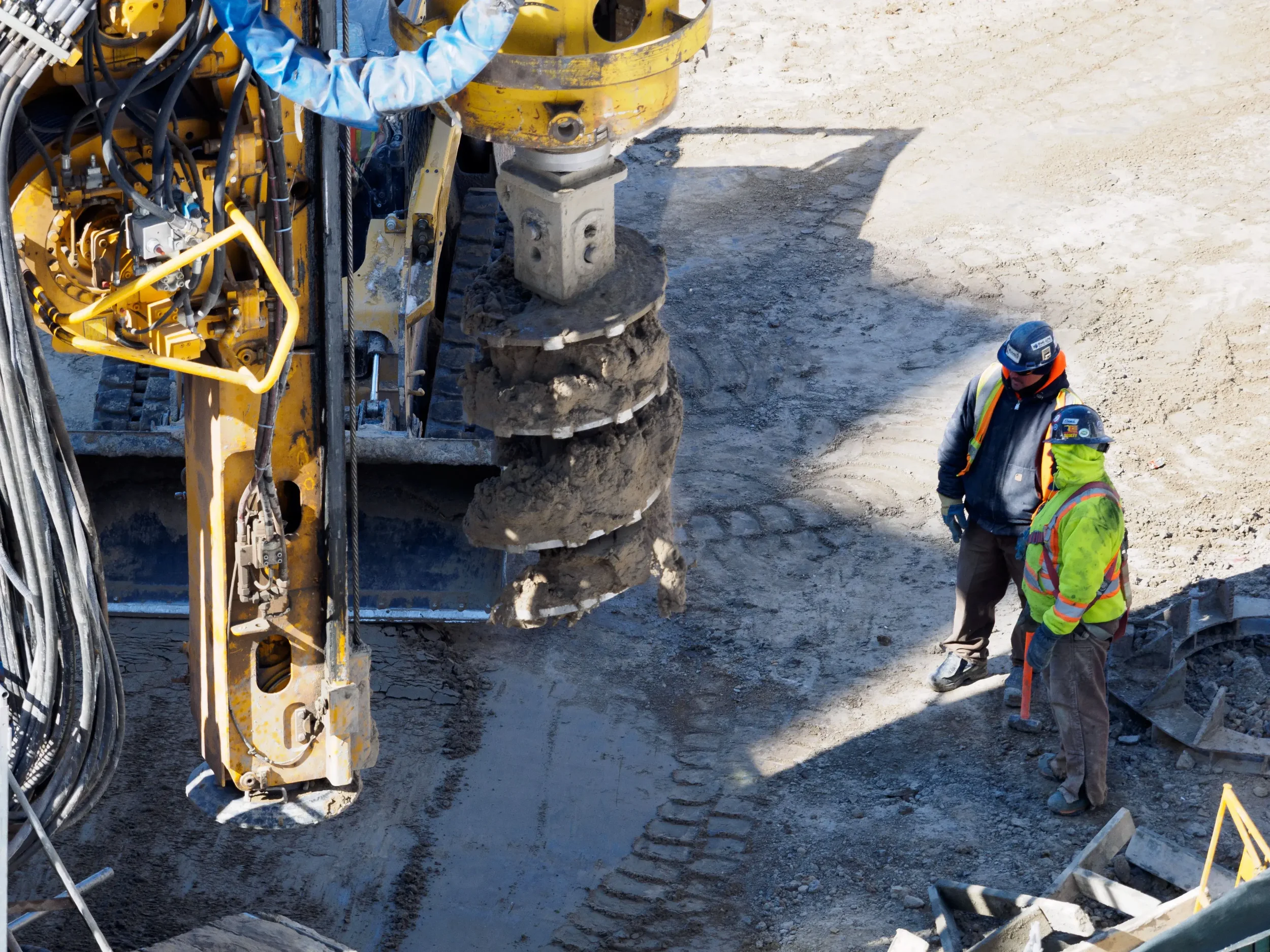 Two Machine Operators in High Vis Jackets Analyze an Industrial Auger Attachment, Potentially Looking for Signs of Overgreased Bearings.