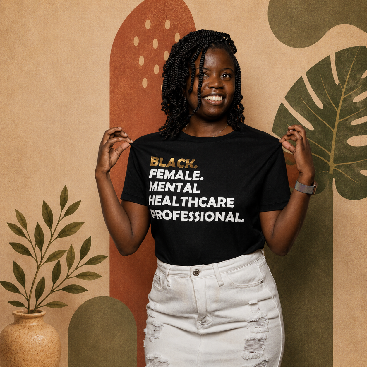 A woman with natural curly hair smiling and holding up a black t-shirt that reads "BLACK. FEMALE. MENTAL HEALTHCARE PROFESSIONAL." in front of a colorful, abstract background with plants.