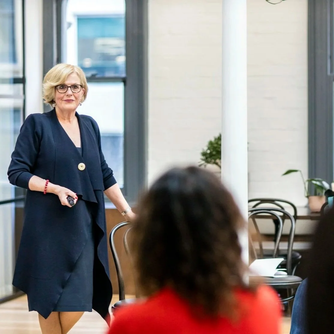 A woman in business attire, holding a remote, standing and speaking in a room with large windows and potted plants, during a small seminar or meeting.