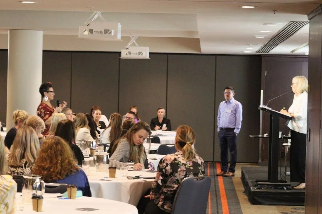 A conference or seminar with a speaker at a podium, people seated at round tables, and a participant with a microphone asking a question.