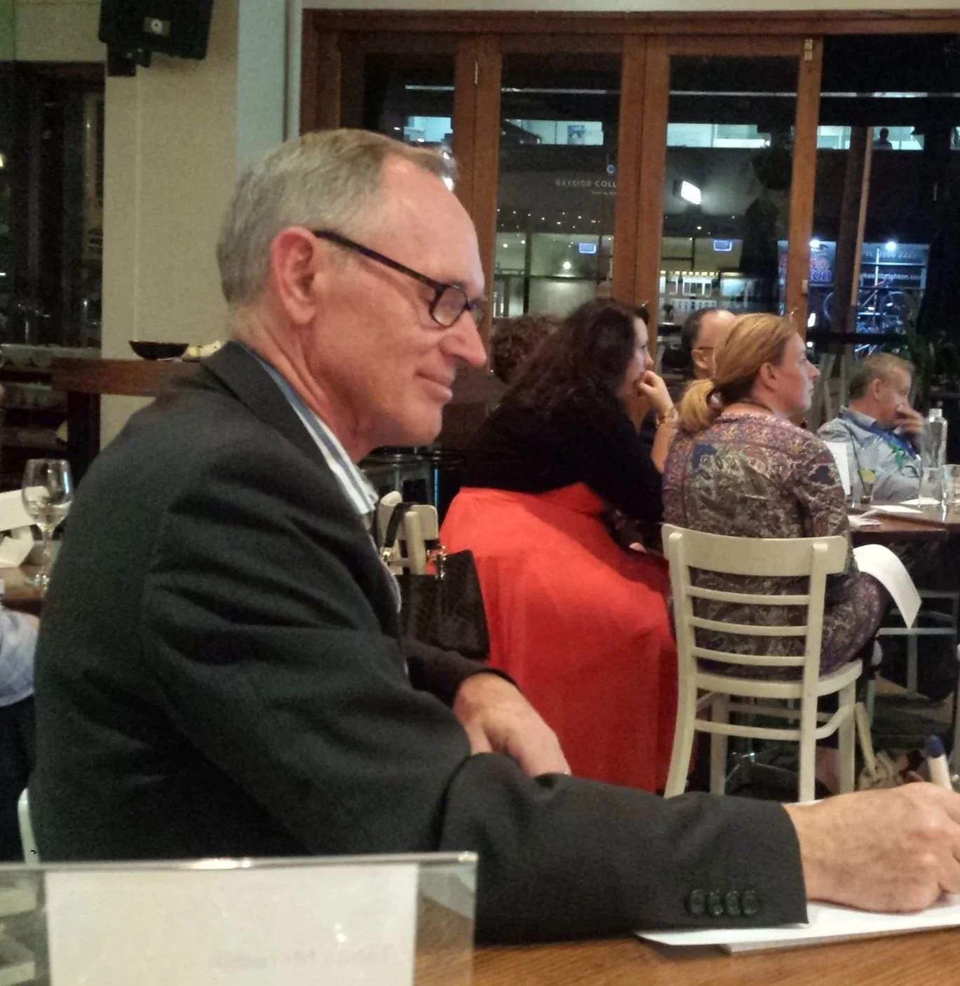 A group of people sitting indoors, attentively listening or participating in an event. Tables with wine glasses are visible around them.