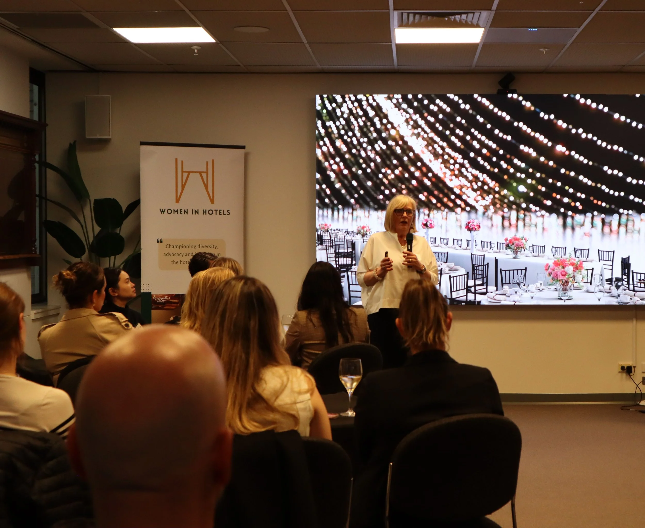 A woman with glasses and a white shirt speaking to an audience at an indoor event, with a large digital screen behind her displaying string lights over an outdoor dining scene, and a standing banner with the logo and text 'Women in Hotels'.