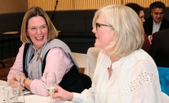 Two women smiling and talking at a table with glasses of wine in a social setting.