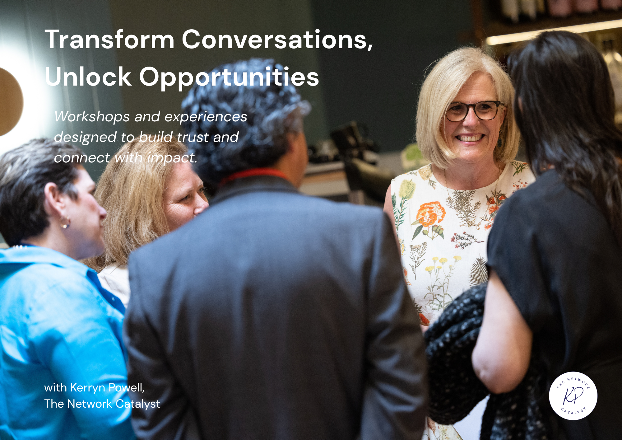 Group of diverse people engaged in conversation indoors, with smiling woman wearing glasses and floral dress at the center.