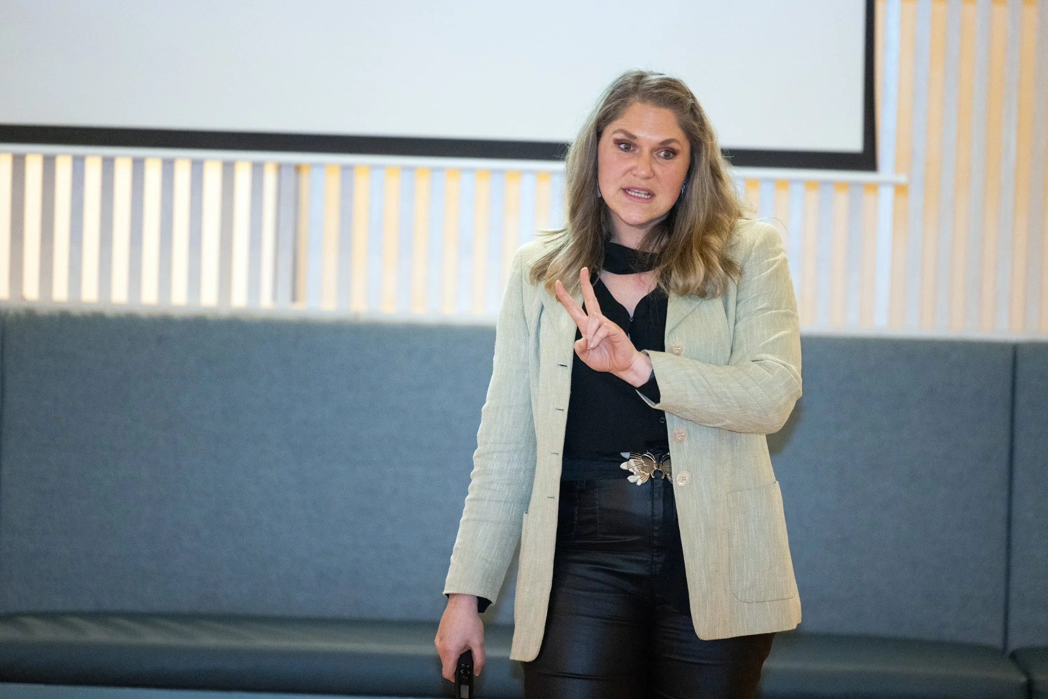 Woman giving a presentation, wearing a light blazer and pointing with two fingers, hardwood panel background.