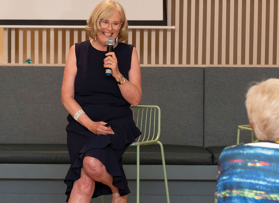 A woman with blonde hair, glasses, and a black dress sitting on a chair, holding a microphone and smiling during a presentation or talk.