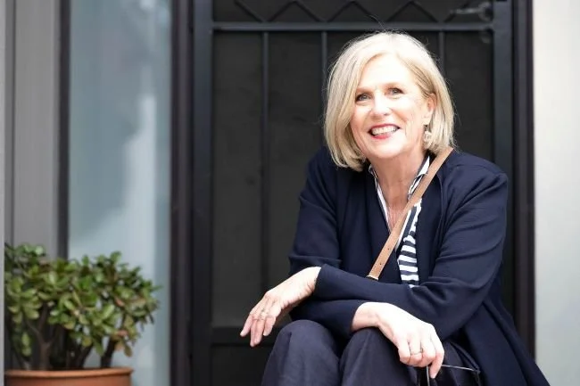 A mature woman with blonde hair, smiling, sitting outside in front of a black gate, wearing a dark blazer and striped shirt.