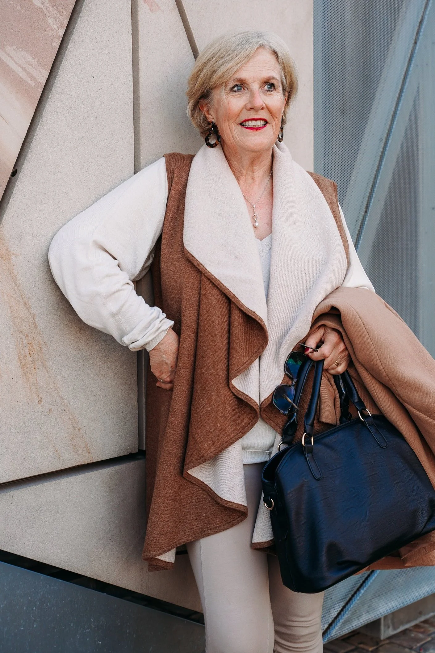 Elderly woman in stylish outfit with brown shawl, beige sweater, and black handbag standing against a modern architectural background.