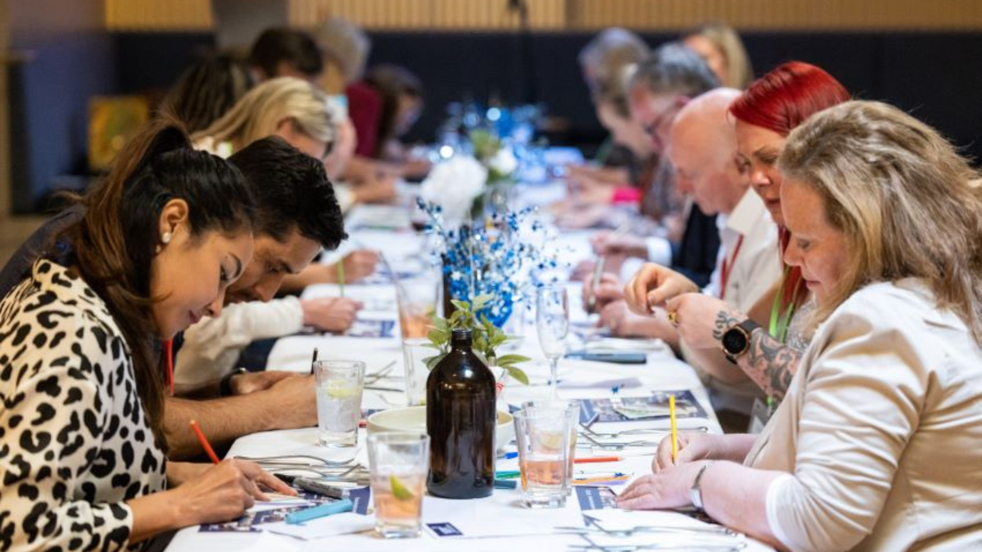 People seated at a long table engaging in a writing activity during a conference or workshop, with blue floral table decorations and various beverages.