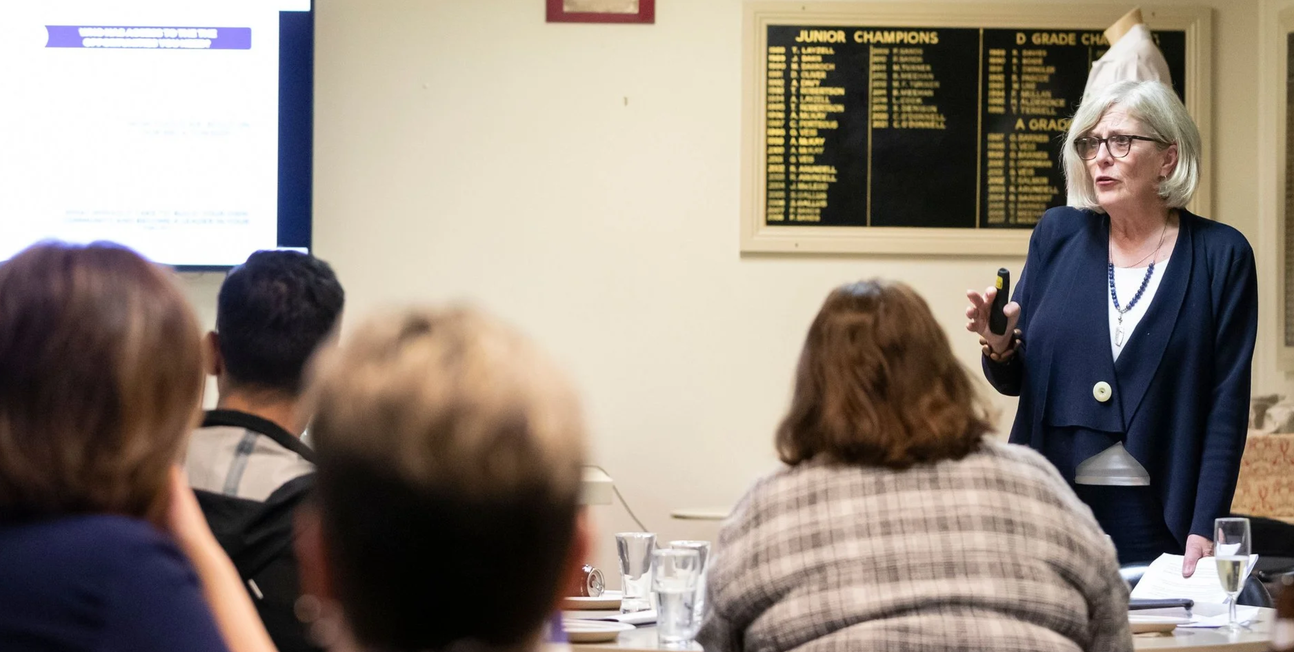 A woman presenting in a room with a small audience. She is holding a remote and speaking to the group, with a presentation slide on a screen behind her and a framed list of achievements on the wall.